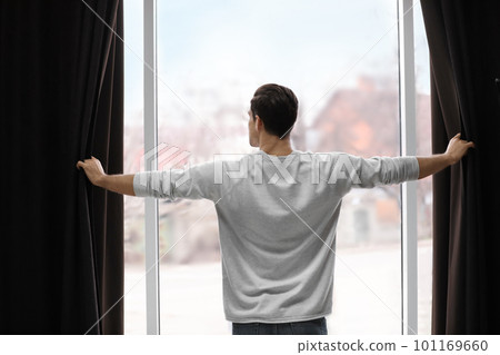 Man opening window curtains at home, back view 101169660