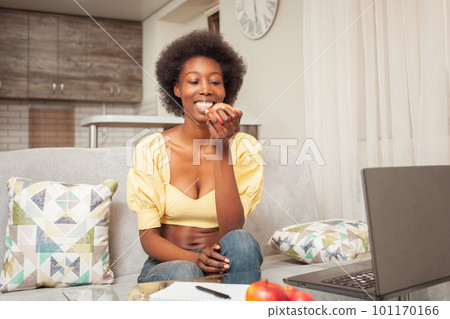 African Black American woman sits in front of laptop, freelance work at home. Snacking, healthy eating, breakfast, diet and weight loss. woman eats an apple fruit. smiles happy 101170166