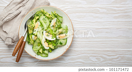 Healthy green avocado salad bowl with boiled eggs, sliced cucumbers, edamame beans, olive oil and herbs on ceramic plate top view, white wooden rustic table background. Copy space 101171472