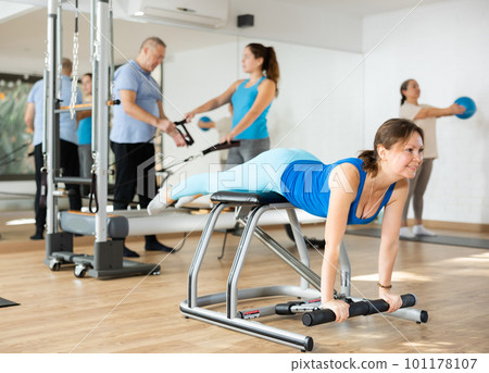 Peaceful middle-aged female in activewear concentrating on stretching exercise on Pilates chair equipment in gym 101178107