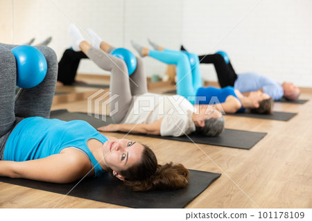 People of different ages pressing core and clutching mini Pilates balls between legs during Pilates training together 101178109