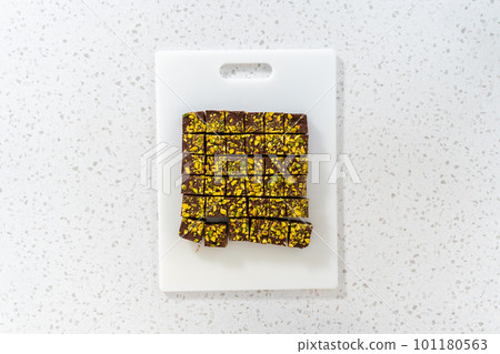 Flat lay. Cutting chocolate pistachio fudge with a large kitchen knife into square pieces on a white cutting board. Flat lay. Cutting chocolate pistachio fudge with a large kitchen knife into square pieces on a white cutting board. 101180563