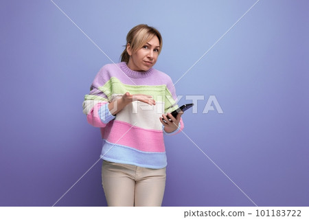 blond doubtful young woman in casual outfit holding a smartphone in her hands on a purple background 101183722