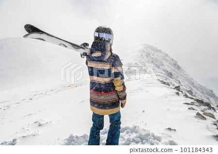 The skier stands on the top of the mountain and enjoys the view of the beautiful winter mountains on a cloudy day. Woman in ski suit holding skis on her shoulder 101184343
