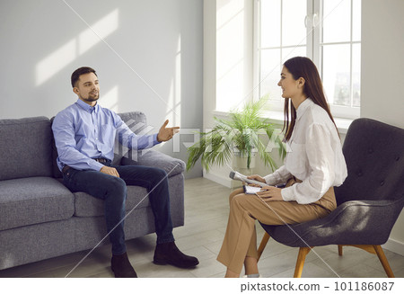 Woman journalist with a microphone interviewing attractive young man sitting on the couch indoors. Woman journalist with a microphone interviewing attractive young man sitting on the couch indoors. 101186087