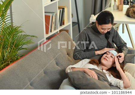 Shot of caring Asian woman taking care of sick girlfriend with fever on couch at home. LGBT, equal rights and love concept Shot of caring Asian woman taking care of sick girlfriend with fever on couch at home. LGBT, equal rights and love concept 101186944