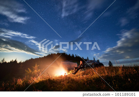 Camping in mountains under evening cloudy sky. Young man having a rest next to campfire, near tourist tent and off-road vehicle on hill. Travelling man sitting and stargazing at night. Camping in mountains under evening cloudy sky. Young man having a rest next to campfire, near tourist tent and off-road vehicle on hill. Travelling man sitting and stargazing at night. 101189101