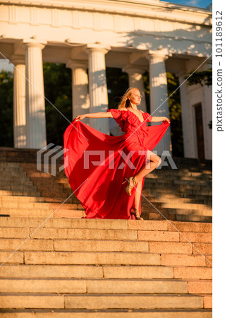 A woman in a long red dress against the backdrop of sunrise, bright golden light of the sun's rays. The concept of femininity, harmony. 101189612