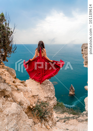 A girl with loose hair in a long red dress descends the stairs between the yellow rocks overlooking the sea. A rock can be seen in the sea. Sunny path on the sea from the rising sun 101189614