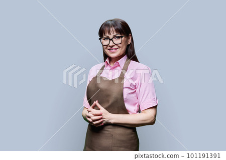 Positive smiling mature woman in apron on gray studio background 101191391