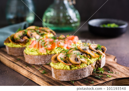 Sandwiches with avocado, fried champignons, tomatoes and herbs on rye bread on a board 101193548