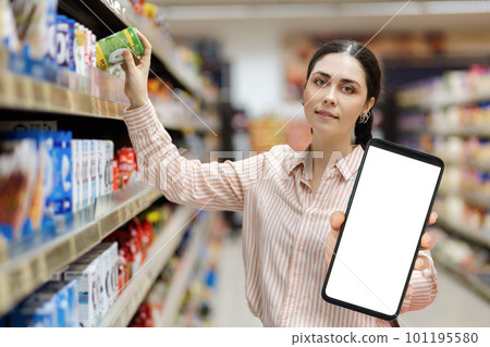 Sales in supermarket. Portrait of young Caucasian pretty woman shows big smartphone with white screen and mock up and takes product from shelf. Concept of discount apps Sales in supermarket. Portrait of young Caucasian pretty woman shows big smartphone with white screen and mock up and takes product from shelf. Concept of discount apps 101195580