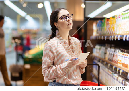 Young Caucasian woman wearing eyeglasses holds product list and choosing from grocery store's assortment. Concept of shopping in supermarket and consumerism Young Caucasian woman wearing eyeglasses holds product list and choosing from grocery store's assortment. Concept of shopping in supermarket and consumerism 101195581