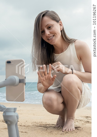 A young female travel blogger greets her viewers with her hand, taking pictures of herself in a blog on a smartphone on the beach by the sea, vertical photo A young female travel blogger greets her viewers with her hand, taking pictures of herself in a blog on a smartphone on the beach by the sea, vertical photo 101196267
