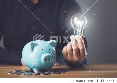 Business, Financial and money planning, Clever thinking about money saving for future and retirement, Man holding light bulb with coin pile and adding coin to piggy bank on the table. Business, Financial and money planning, Clever thinking about money saving for future and retirement, Man holding light bulb with coin pile and adding coin to piggy bank on the table. 101197271