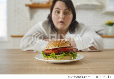 Young fat excited woman sitting at the table in kitchen looking at the plate with big burger. 101198015