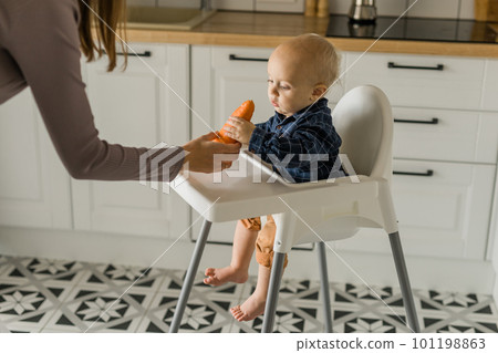 Baby boy sitting in baby chair eating carrot on kitchen background copy space - baby feeding concept 101198863