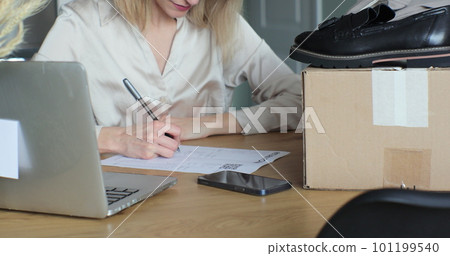 Young woman filling out a paper form, wanting to return shoes. Female writes a return order and sits at a table with a laptop at home. 101199540