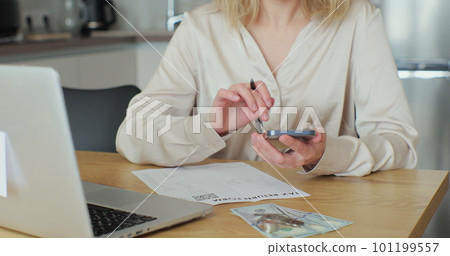 Close up woman fills out paper document and counts on the phone screen. Young woman is writing tax return form and looking at laptop at table in home room. 101199557