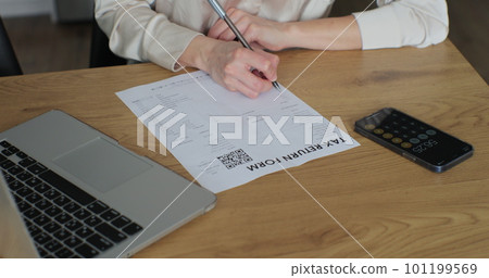 Young woman is writing tax return form and looking at laptop at table in home room. Close up woman fills out paper document and counts on the phone screen. Young woman is writing tax return form and looking at laptop at table in home room. Close up woman fills out paper document and counts on the phone screen. 101199569