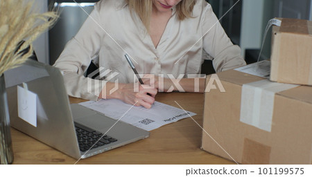A young woman writes a return order and sits at a table with a laptop at home. Close-up video of a female customer writing on paper and filling out a form, wanting to return a product. 101199575