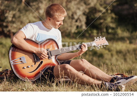 Attractive European male teenager sits on grass, plays acoustic guitar, recreats during summer day, practices new song, sings, enjoy calm atmosphere outside. People and entertainment concept 101200554