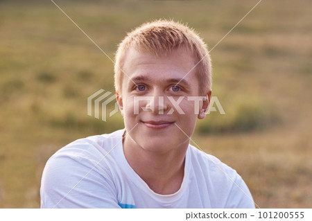 Horizontal shot blue eyed light haired teenager dressed in casual white t shirt, has genstle friendly smile, poses outdoor against blurred background, enjoys peaceful atmosphere. Lifestyle concept Horizontal shot blue eyed light haired teenager dressed in casual white t shirt, has genstle friendly smile, poses outdoor against blurred background, enjoys peaceful atmosphere. Lifestyle concept 101200555
