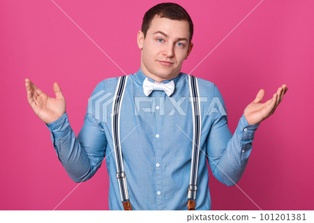 Close up portrait of careless guy spreading hands in helpless gesture, young man wearing blue shirt, suspenders and white bowtie shrugging shoulders, posing isolated over pink studio background. Close up portrait of careless guy spreading hands in helpless gesture, young man wearing blue shirt, suspenders and white bowtie shrugging shoulders, posing isolated over pink studio background. 101201381