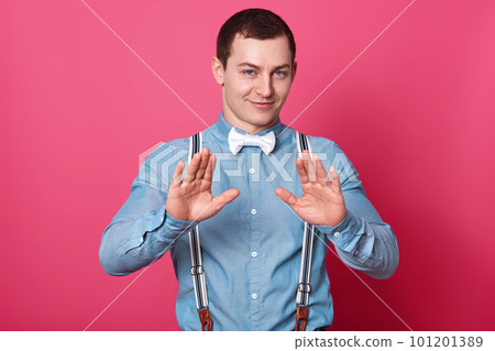 Young attractive male with short hairstyle. Model raises palms and shows stop sign, wears blue shirt with suspenders and white bow tie, isolated over pink concrete wall. Body language concept. Young attractive male with short hairstyle. Model raises palms and shows stop sign, wears blue shirt with suspenders and white bow tie, isolated over pink concrete wall. Body language concept. 101201389