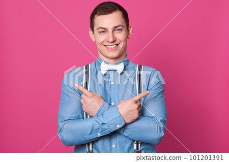 Indoor shot of happy funny young man laughing and looks excited, stands with crossing hands pointing fingers aside at copy space for advertisment or promotion isolated over pink studio background. Indoor shot of happy funny young man laughing and looks excited, stands with crossing hands pointing fingers aside at copy space for advertisment or promotion isolated over pink studio background. 101201391