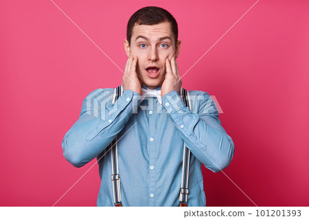Surprised young Caucasian man with raised eyebrows and open mouth, dressed blue shirt, white bow tie and suspenders, stands against pink background, has astonished expressions, keeps hands on cheeks. 101201393