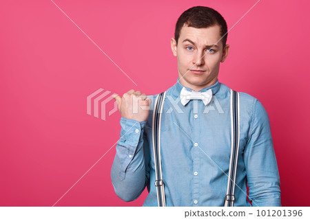 Studio shot of attractive male in blue shirt, suspenders and white bow tie, shows with thumb finger aside, isolated over pink studio background. Copy space for advertisment and promotion text. 101201396