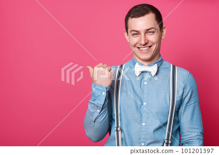 Happy young handsome man in shirt, suspenders and bow tie, points away with thumb finger and smiling while standing against pink background, suggests use copy space for logo, has funny expression. 101201397