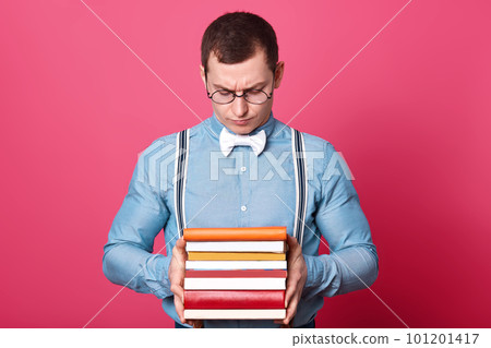 Indoor studio shot of dark haired confused model posing isolated over bright pink background, holding books in his strong hands, looking at them, reading text and assignments. Youth and study concept. 101201417