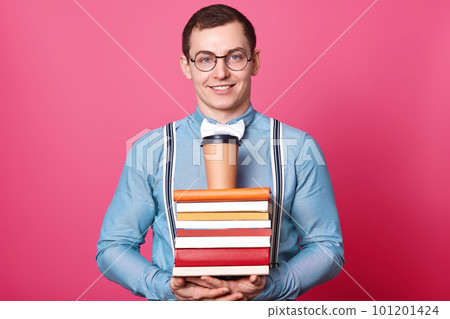 Close up portrait of young student in blue shirt in one tone, holds coffee on huge stack of books, wants to have rest and drink something bracing, isolated on rosy background. Education concept. 101201424