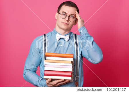 Image fo young student man holding stack of books and having headache, keeps hand on head, model poses on rose studio background, looks directly at camera. Copy space for your text. Education concept. Image fo young student man holding stack of books and having headache, keeps hand on head, model poses on rose studio background, looks directly at camera. Copy space for your text. Education concept. 101201427