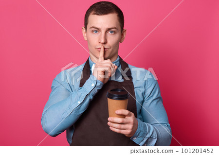 Dont tell secret of making coffee. Mysterious handsome male barista works in cafe, makes hush gesture, keeps fore finger over mouth, holds disposable cup of hot beverage, dressed in apron poses indoor 101201452