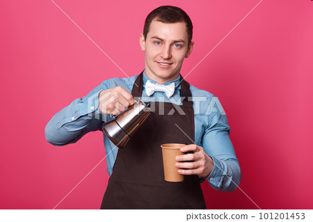 Portrait of professional male barista pours aromatic coffee into paper cup, wears blue shirt, white bow tie and brown apron, isolated over pink background. Young handsome man works in coffe shop. 101201453