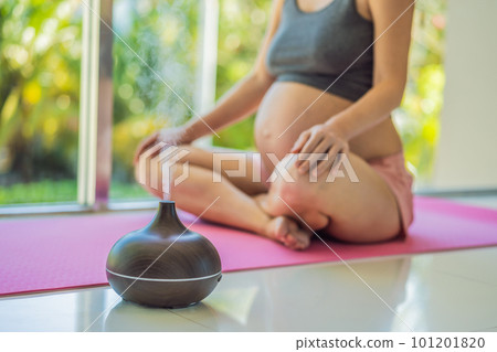 Young pregnant woman doing yoga exercises and meditating at home with an aroma diffuser. Health care, mindfulness, relaxation and wellness concept. Aromatherapy during pregnancy 101201820