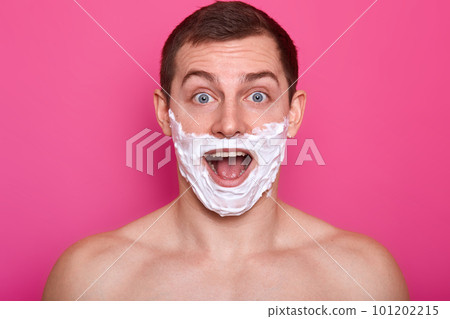 Studio shot of cheerful surprised man with shaving foam on his face looks directly at camera with open mouth, has astonished facial expression, looks happy, model posing isolated over pink background. Studio shot of cheerful surprised man with shaving foam on his face looks directly at camera with open mouth, has astonished facial expression, looks happy, model posing isolated over pink background. 101202215