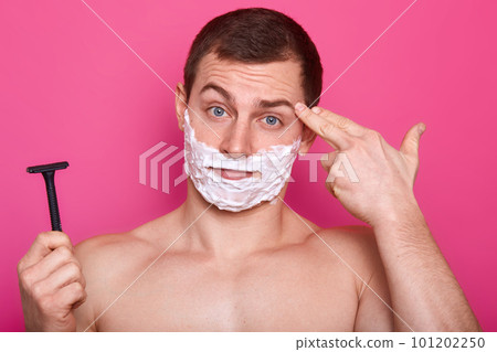 Close up portrait of handsome man with shaving foam on face, keeps razor in hand ready for shaving, pretends killing himself, shoots in temple with hand, looks unhappy, shows suicide gesture. Close up portrait of handsome man with shaving foam on face, keeps razor in hand ready for shaving, pretends killing himself, shoots in temple with hand, looks unhappy, shows suicide gesture. 101202250