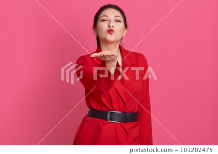 Indoor portrait of lovely charming pretty brunette sending air kiss, raising one hand, making gesture, looking directly at camera, wearing stylish belt and red jacket. People and emotions concept. Indoor portrait of lovely charming pretty brunette sending air kiss, raising one hand, making gesture, looking directly at camera, wearing stylish belt and red jacket. People and emotions concept. 101202475