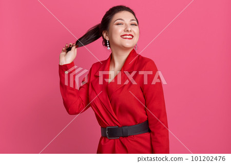 Picture of cheerful slim magnetic female smiling sincerely, having beautiful smile, holding her ponytail, wearing red jacket and black belt, being in high spirits. People and emotions concept. Picture of cheerful slim magnetic female smiling sincerely, having beautiful smile, holding her ponytail, wearing red jacket and black belt, being in high spirits. People and emotions concept. 101202476