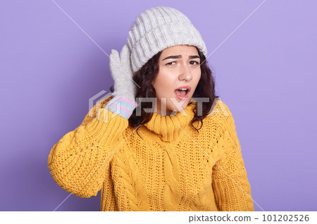 Image of curly haired emotional energetic young woman standing isolated over lilac background in studio, opening yellow sweater and hat, gloves, putting hand to ear, trying to hear something. 101202526