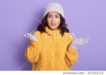 Portrait of unsatisfied shocked brunette raising both hands, making gestures, looking directly at camera, wearing white hat, gloves and yellow sweater, standing isolated over lilac background. 101202532