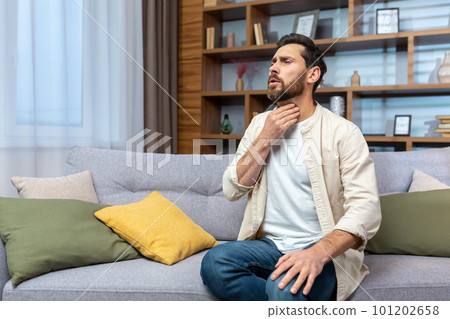 Sore throat. A young man sits at home on the sofa and holds his throat, feels a sore throat, burning sensation, discomfort. Sore throat. A young man sits at home on the sofa and holds his throat, feels a sore throat, burning sensation, discomfort. 101202658