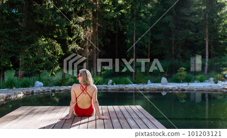 Rear view of young woman in swimsuit sitting on pier by lake during summer vacation in mountains. 101203121