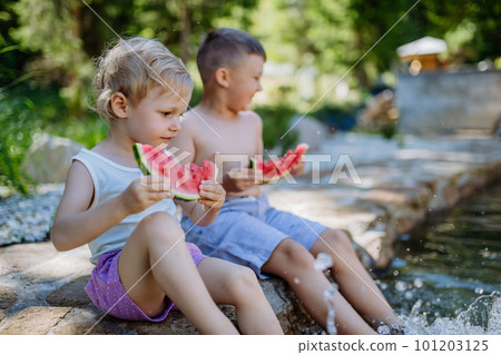 Little chidren sitting near lake and eating watermelon on hot sunny day during summer vacation. 101203125