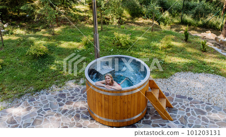 Young woman enjoying wooden bathtub with a fireplace to burn wood and heat water in backyard in mountains. High angle view. Young woman enjoying wooden bathtub with a fireplace to burn wood and heat water in backyard in mountains. High angle view. 101203131