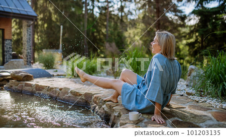 Happy young woman sitting by pond near cottege and enjoying cup of morning coffee on summer vacation in mountains. High angle view. 101203135